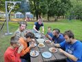 uczniowie siedzą przy stole na dworze przy ognisku jedzą kiełbasy kaszanki tosty nauczyciel pomaga kroic uczniowi kiełbasę