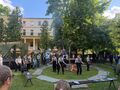 zaproszeni goście i harcerze wspólnie śpiewają i wykonują pląsy stoją w kole wokół ogniska w parku w tle budynek szkołyjpg