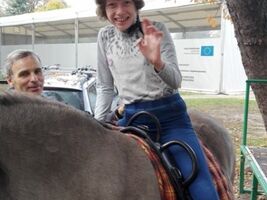 dziewczynka siedzi na grzbiecie konia o imieniu Hejnał, unosi rękę w kierunku obiektywu aparatu, obok asekuruje hipoterapeuta